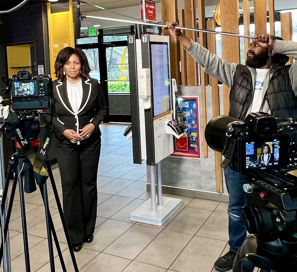 A woman in a formal outfit stands for an interview in a fast-food restaurant, while a man adjusts a camera light stand nearby.