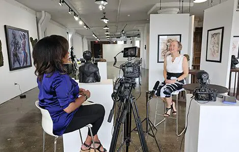 Two women seated having an interview in an art gallery with cameras and lighting equipment.