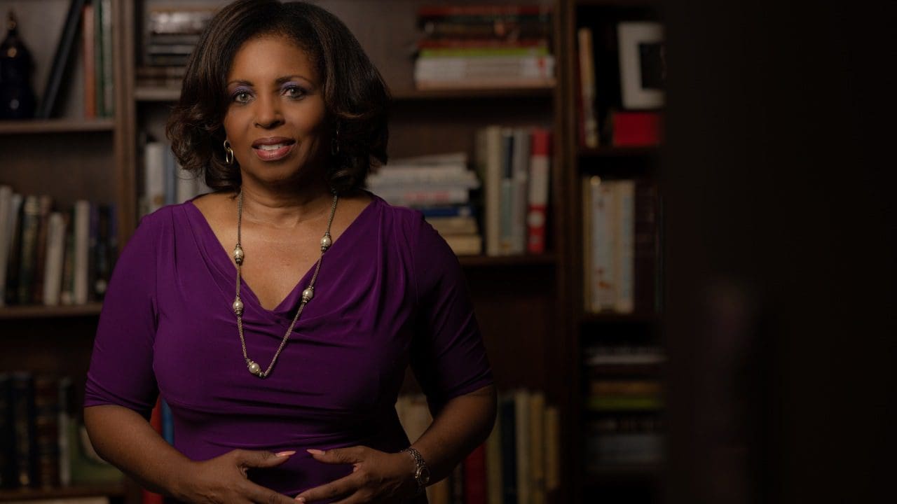 African-american woman in a purple top and necklace standing confidently in front of a bookshelf, softly lit with a focus on her.