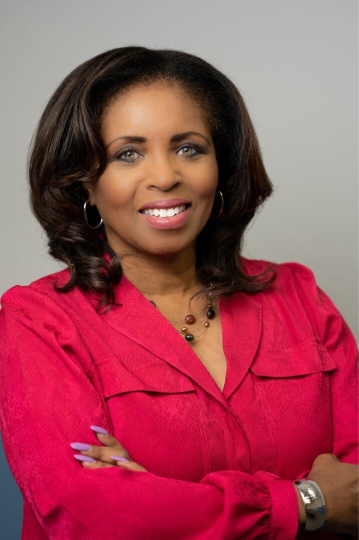 A professional portrait of a woman in a red blouse smiling confidently with crossed arms against a gray background.