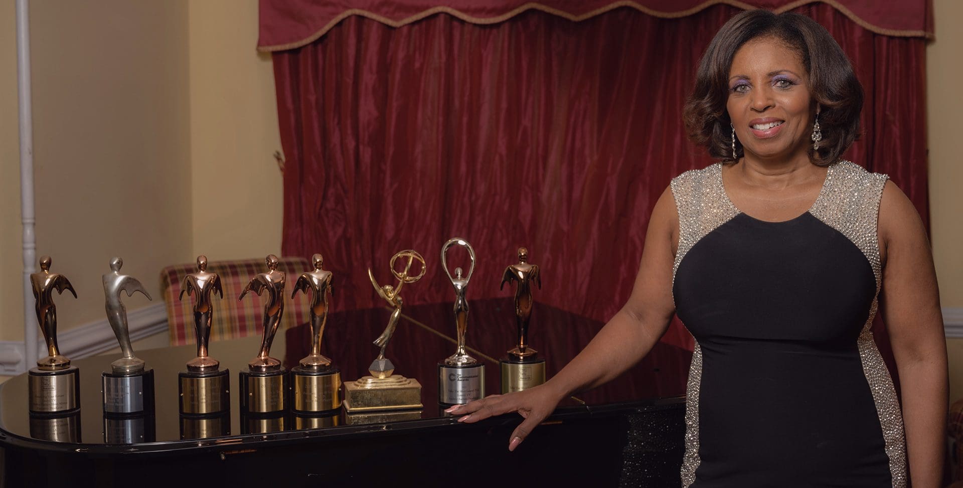 A woman stands next to a table displaying a collection of various awards, including trophies and plaques, in an elegant room with a red curtain backdrop.