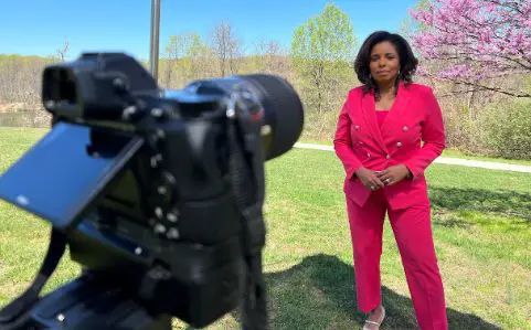 A woman in a pink blazer stands with crossed arms, smiling, with blooming cherry trees in the background.