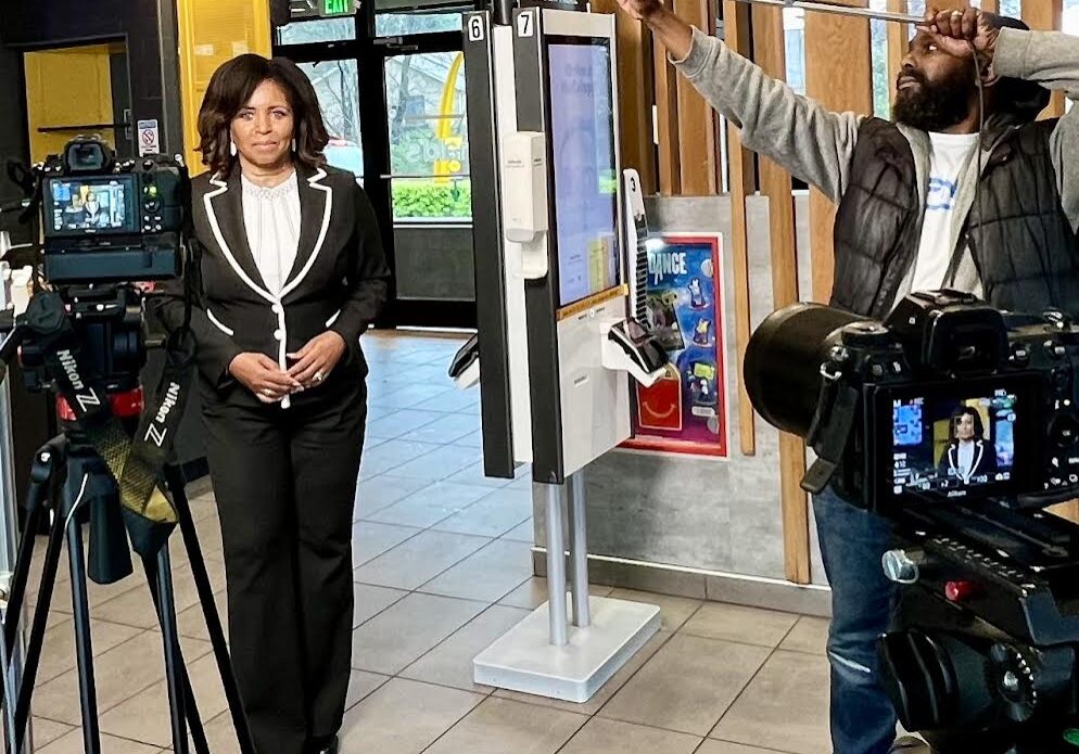 A woman in a formal outfit stands for an interview in a fast-food restaurant, while a man adjusts a camera light stand nearby.